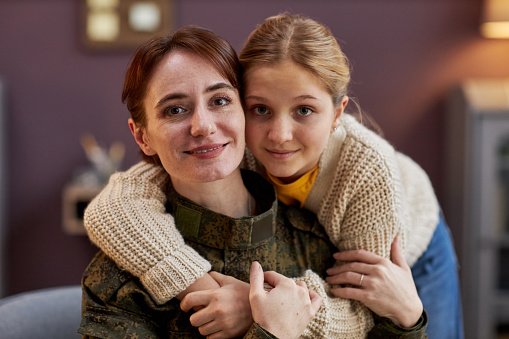Portrait of smiling military woman with daughter embracing and looking at camera at home