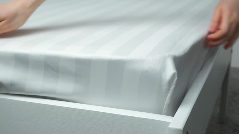 A white satin sheet with an elastic band is put on the mattress of the bed, a close-up of the hands. Bedclothes.