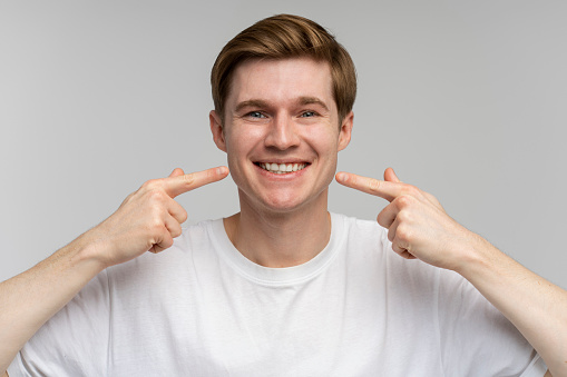 Portrait of happy cheerful man points index fingers at smile shows white teeth, looking at camera. Indoor studio shot isolated on white background
