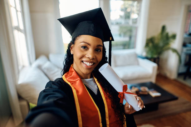 Happy black female student holding university diploma while taking selfie at home.