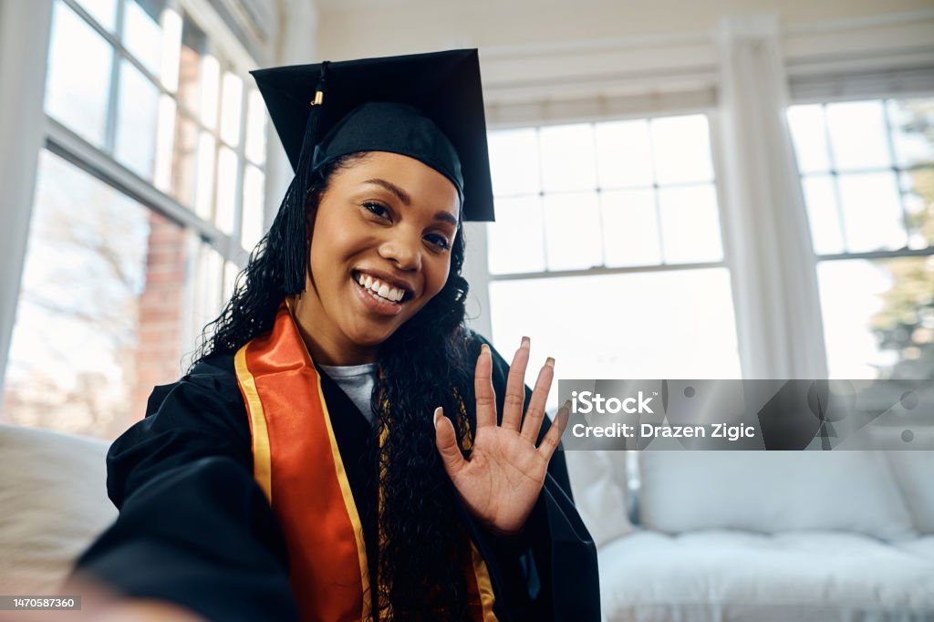 Happy black graduate student  waving during video call from home. Happy African American woman in graduation gown taking selfie at home. Graduation Stock Photo