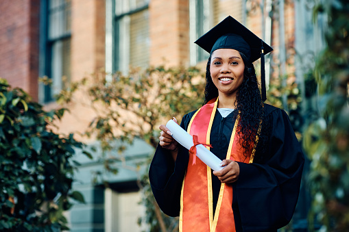 A black young woman, 18 years old, standing outside a school building wearing a graduation cap and gown, holding a diploma.