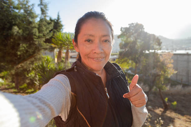 femme adulte souriant à l’appareil photo avec le pouce levé tout en prenant un selfie - femme hispanique prenant une photo avec son téléphone portable - femme latine profitant du coucher de soleil - women poverty senior adult mature adult photos et images de collection