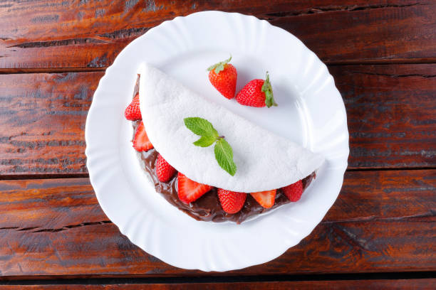 homemade tapioca or beiju stuffed with strawberry and chocolate on white plate over rustic wooden table stock photo
