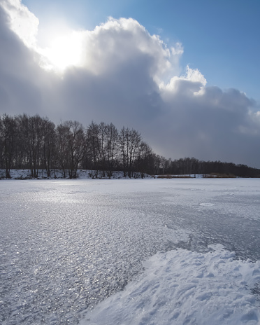 Winter landscape with a frozen steppe river, trees on the banks and low clouds through which the sun breaks through, a winter view of a frozen river