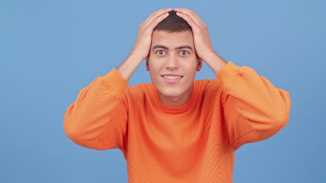 Wow, shock and surprise, portrait of man with hands on head shocked by news or announcement of discount sale in studio. Gen z person with surprised face, emoji or meme isolated on blue background.