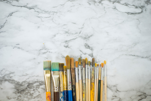 Small group of used artists paintbrushes of various sizes and shapes lined up together in an uneven row on a white marble counter top background.