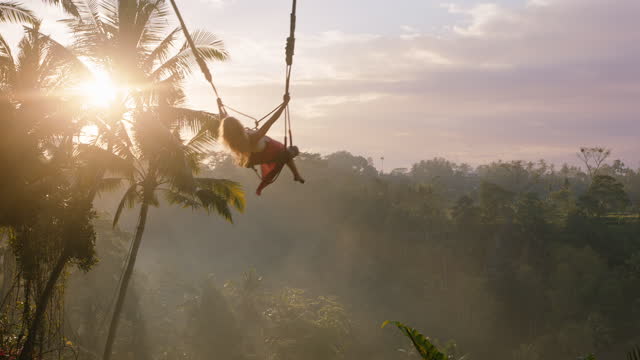 happy woman swinging over jungle at sunrise travel girl enjoying exotic vacation on swing with sun flare shining through palm trees in tropical rainforest holiday lifestyle freedom