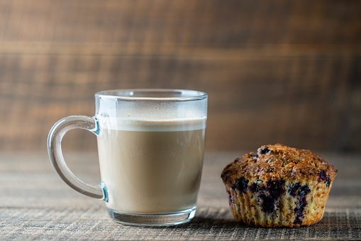 Delicious muffin with blueberries and cappuccino glass cup on a wooden table, close up. Fresh cupcake and coffee for breakfast