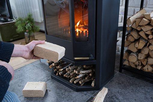 Hands kindle the hearth with economical briquettes. Fuel briquettes made of pressed sawdust for kindling the furnace - economical alternative eco-friendly fuel for the fireplace in the house.