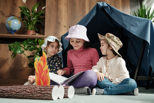 Two child girls and one toddler boy playing camping game at home and reading book together, concept for activities for activities at home for children