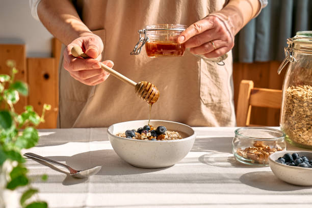 mulher preparando dieta saudável vegano café da manhã nutritivo. mão feminina derramando mel na tigela com mingau de aveia com nozes e mirtilos. - english walnut - fotografias e filmes do acervo