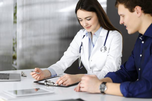 Friendly young woman-doctor is listening to her patient, while they are sitting together at the desk in a cabinet. Physician is holding a clipboard in her hands for filling up medication recommendations. Medicine and healthcare concepts. una amistosa joven-doctora está escuchando a su paciente, mientras están sentados juntos en el escritorio en un gabinete. la médica sostiene un portapapeles en sus manos para completar las recomendaciones de medicamentos. conceptos de medicina y asisten