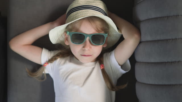 Sad little girl in sunglasses with green rim lies on sofa