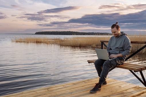 A handsome young man is resting and working on a laptop near the sea. A freelancer works sitting on a wooden bench on a pier by the water.
