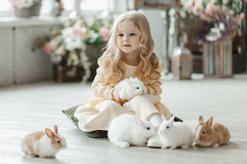 A little smiling girl in a yellow dress is playing on the floor in a room with rabbits. A baby and a rabbit. The living room is decorated for Easter