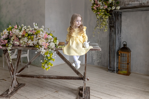 A little smiling girl in a yellow dress is sitting with rabbits on a wooden table with flowers.A baby and a rabbit. The concept of Easter