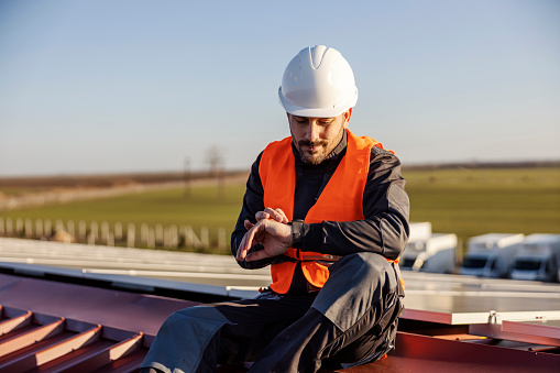 A worker in protective work wear is sitting on the roof and looking at wristwatch. Break is over.