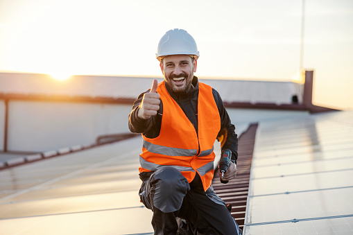The worker approving eco live and solar panels using while crouching on the rooftop with drill in his hands.