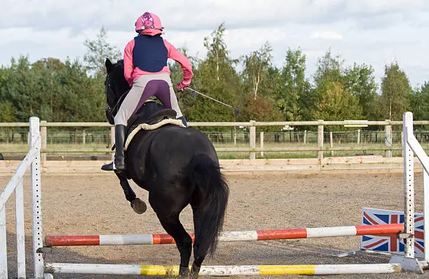Young woman riding over a jump in an outdoor arena, riding away from the camera.