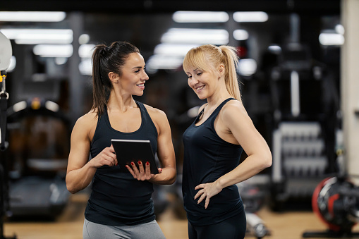 A happy female coach is pointing at the tablet while sportswoman is looking at the progress on tablet in a gym.