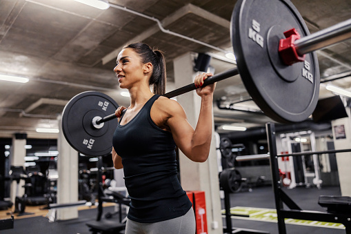A happy muscular female bodybuilder is lifting barbell in a gym and doing exercises for shoulders.