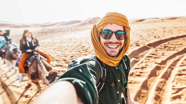 turista feliz que se divierte disfrutando de un paseo en camello en grupo en el desierto - viaje, estilo de vida, actividades de vacaciones y concepto de aventura - morocco desert camel africa fotografías e imágenes de stock