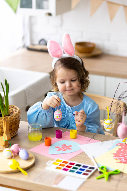 portrait of cute pretty little girl in blue dress and funny rabbit ears making craft, diy eggs for easter holiday home decoration. spring colours, bright flowers. kid's domestic life, hobby, enjoyment - child easter egg home improvement easter imagens e fotografias de stock