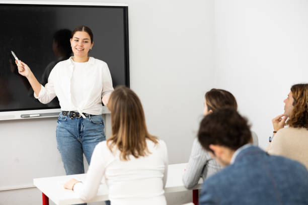 istock Young female business coach explaining topic while standing against group of people sitting at desks in front of her 1470691097