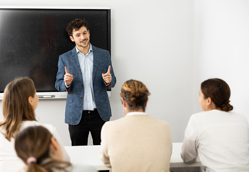 Young male teacher near interactive board, gesturing with hand while consecrating topic of lesson for adults in classroom