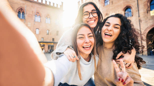 Three young women taking selfie picture with smart mobile phone on city street - Happy beautiful female friends smiling at camera outdoors -  Life style concept with cheerful girls enjoying vacation stock photo