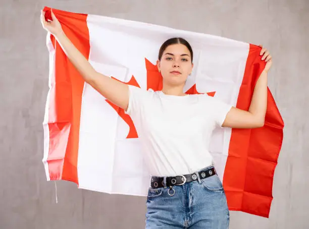 Serious patriotic young woman holding state flag of Canada against gray wall background indoors