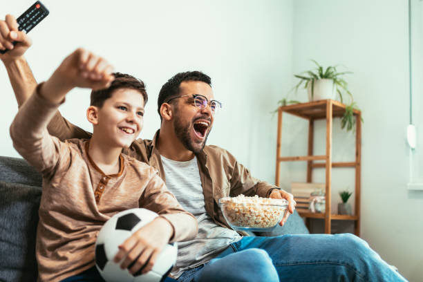 Boy watching soccer match with father at home, sitting on sofa