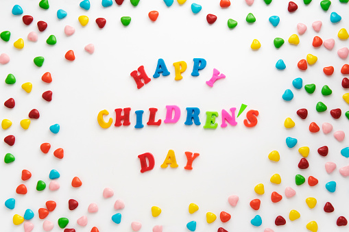 Happy Children's Day. Colorful text surrounded by sweets on white background. Plastic voluminous multi-colored child toy letters, small colored candies in form of hearts. Greeting or invitation postcard for international holiday. Top view. Cartoon flat lay.