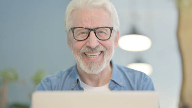 istock Close Up of Old Man Smiling at Camera while using Laptop 1470693509