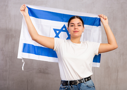 Portrait of serious patriotic young female holding national flag of Israel against gray wall background