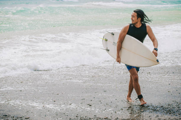 surfista masculino chinês asiático caminhando na praia de volta do mar depois de surfar onda no oceano pacífico - surfing sport extreme sports success - fotografias e filmes do acervo