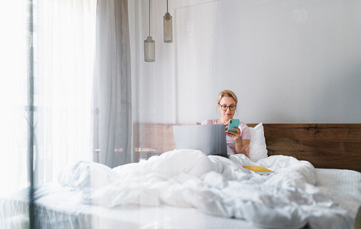 Cheerful woman using laptop and phone in the morning in cozy bed at home. Cute blonde woman working at home