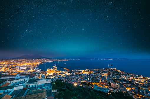 Naples, Italy. Skyline Cityscape In Evening Lighting. Tyrrhenian Sea And Landscape With Volcano Mount Vesuvius. City In Night Illuminations. Bright Blue Night Sky. Amazing Night Starry Sky Background.