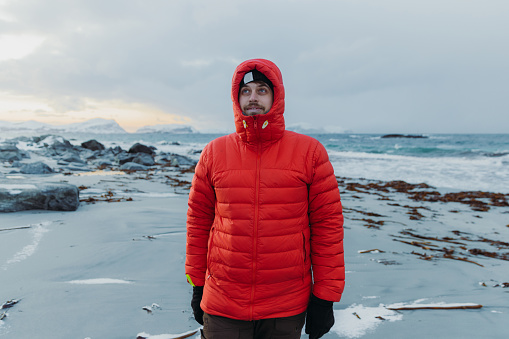 A male in bright colorful winter jacket walking at the seashore with a view of the islands. with snowcapped mountains in Scandinavia