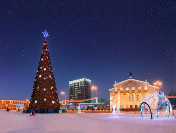 Photo of Gomel, Belarus. Amazing Bright Starry Sky Above Xmas Christmas Tree In Lenin Square And Drama Theatre With Christmas New Year Decorations In Evening Night Illumination. Evening View Of City Center