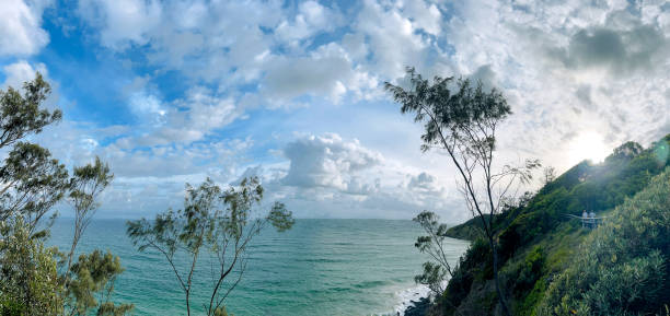 Horizontal high angle landscape panoramic view of the rising sun, the turquoise coloured Pacific Ocean, a blue sky with fluffy white clouds, coastal trees and scrub forest as seen from the Cape Byron walking track, Byron Bay, north coast NSW in Summer.