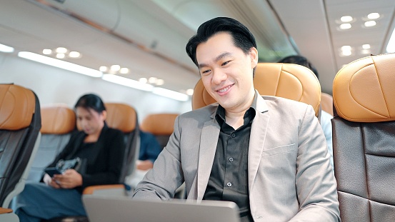 Happy Asian handsome businessman in suit using laptop notebook sitting inside an airplane
