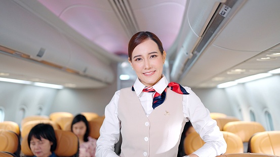 Attractive young Asian stewardess in uniform looking and smiling to the camera in airplane, Cabin crew or air hostess occupation concept