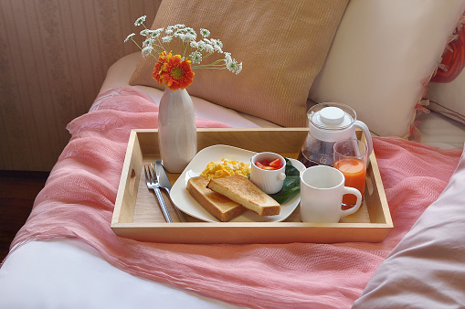 Breakfast in bed with espresso coffee, croissant, honey and jam on the wooden tray, close up. White cup of coffee on the gray bedsheet with pillow and blanket. Happy morning ritual