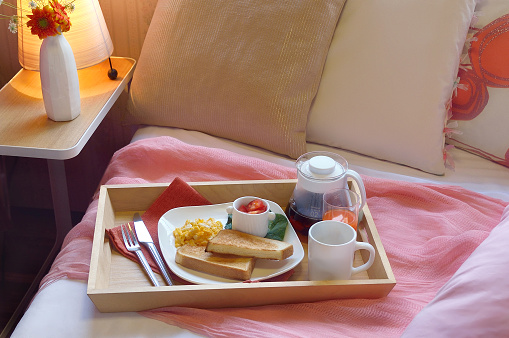 Breakfast on the bed inside a bedroom. Wooden try with coffee, orange juice, croissant, jam ,strawberry and red rose flower. Romantic surprise.