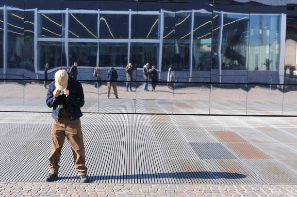 Boy in a cap standing as a model in front of a glass wall with windows with his head bent during a scence shooting act on a sunny day