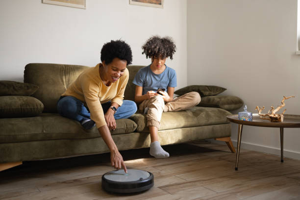 Let's turn on this robotic vacuum cleaner! Happy black single mother tuning on robotic vacuum cleaner while relaxing with her son on sofa in the living room. smart home family stock pictures, royalty-free photos & images