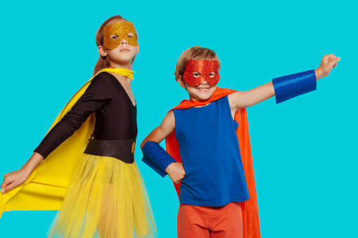 Two blond children wear bright superhero costume, on children party, pretend being supernatural having super powers. Happy childhood concept. Isolated over blue background.