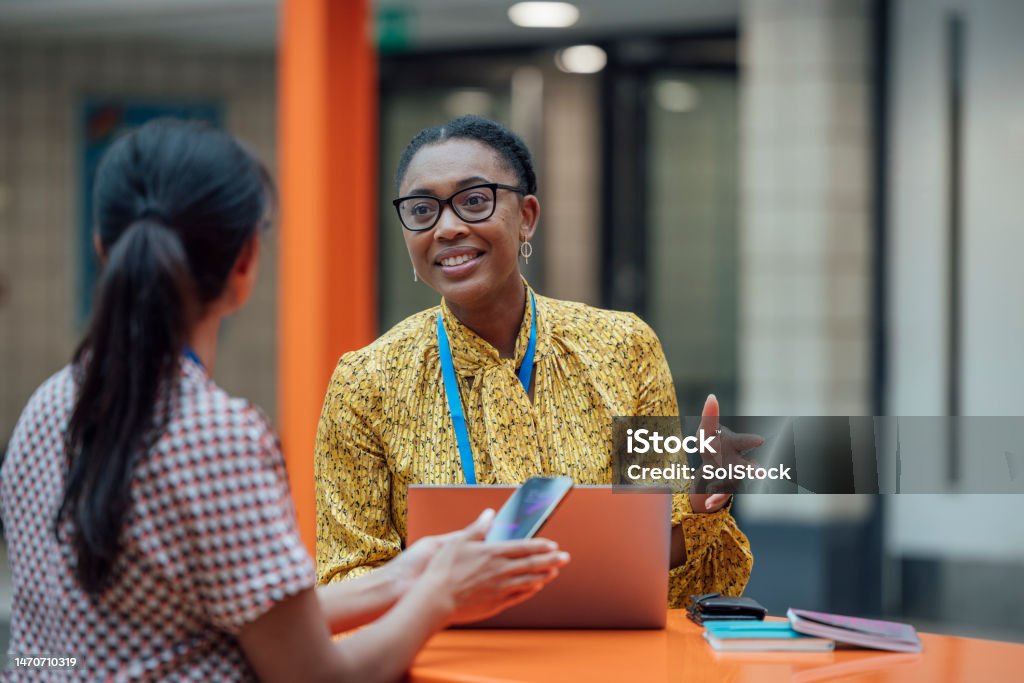 How Was Your Last Lesson? Two female teachers sitting at a table, having a discussion in the lobby area in the school they work at in Gateshead, North East England. They are talking about school issues together while using a laptop. Teacher Stock Photo